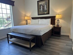 Une chambre avec un grand lit, deux tables de chevet avec des lampes, un banc au pied du lit et une fenêtre donnant sur un paysage verdoyant.