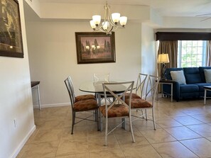 A dining area with a glass table and metal chairs, a chandelier, a painting on the wall, and a sofa in the background. - 4307 Spacious 3/2 Resort Condo Near Disney (Orlando)