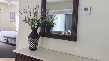 Un meuble en bois foncé, doté de tiroirs et surmonté d’un miroir, est placé contre un mur où se trouvent un thermostat et une fenêtre.