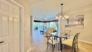 A dining area with a glass table and chairs, a ceiling fan, and a painting on the wall. - 5302 Spacious 3/2 Resort Condo Near Disney (Orlando)