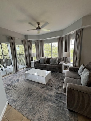Ein geräumiges Wohnzimmer mit Deckenventilator, großem Fenster mit Vorhängen, grauem Sofagarnitur, weißem Couchtisch und Teppich.
