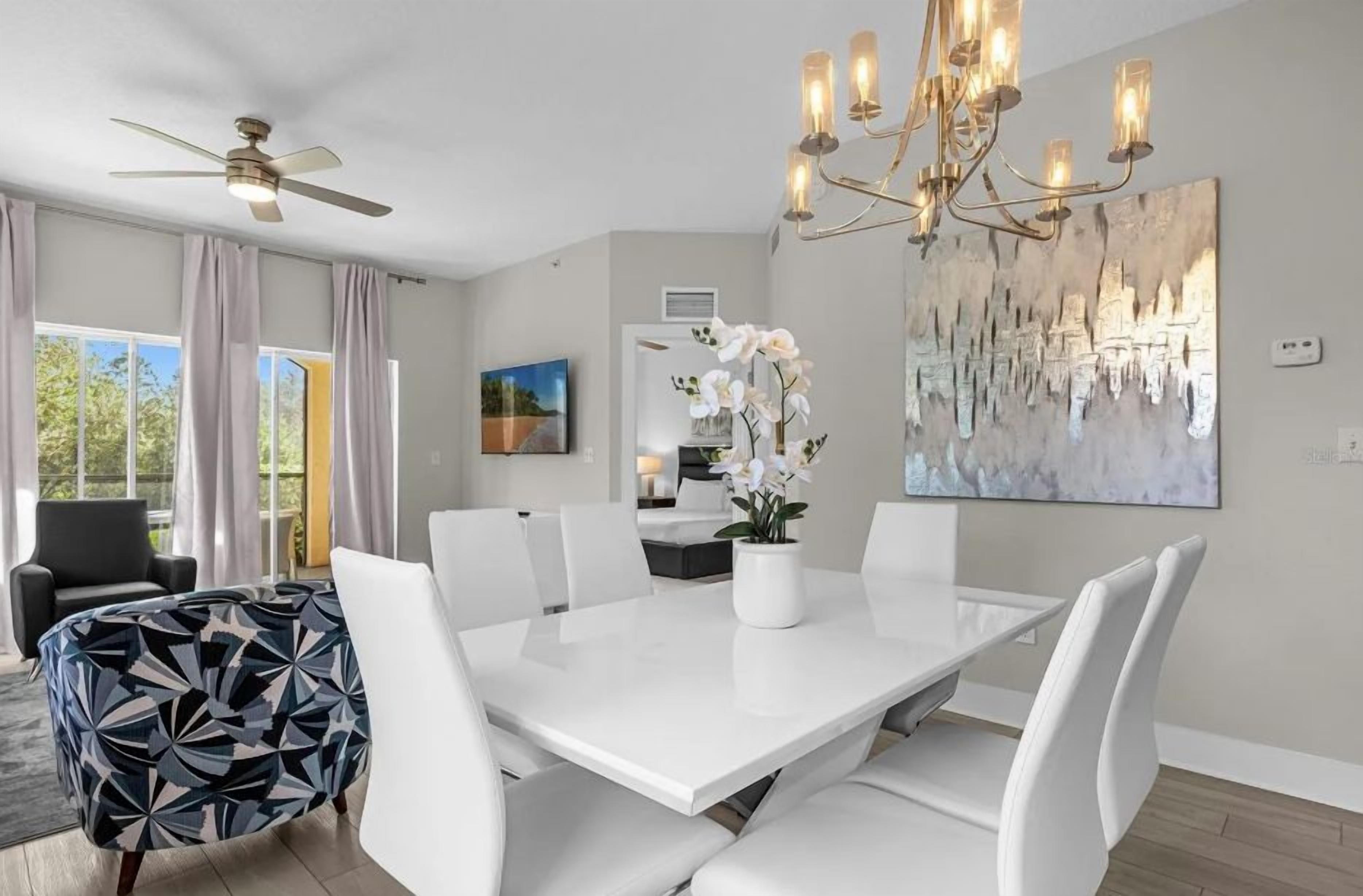 Un espace repas avec une table blanche, des chaises blanches, un ventilateur de plafond et un lustre.