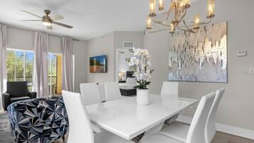 Un espace repas avec une table blanche, des chaises blanches, un ventilateur de plafond et un lustre.