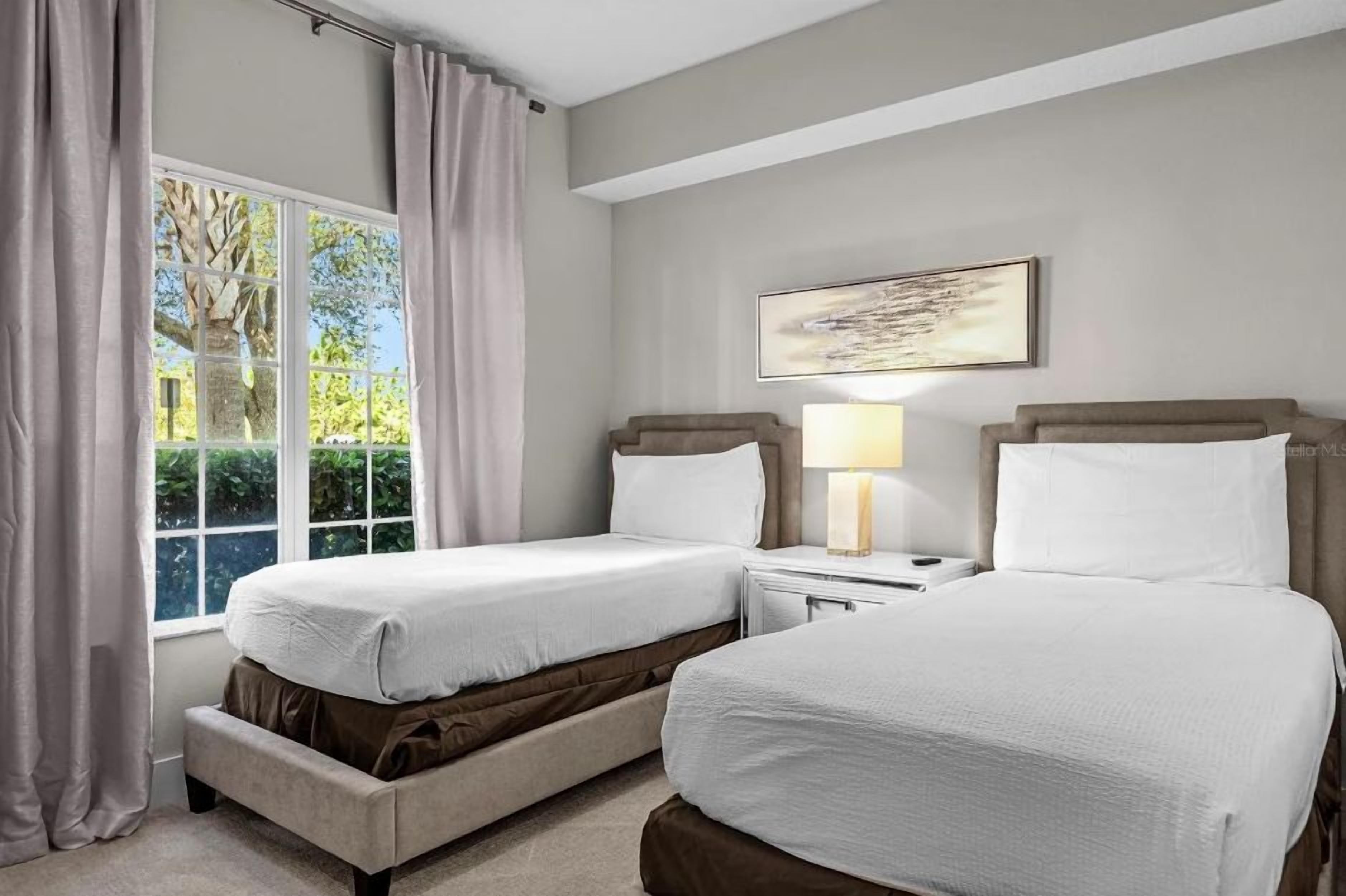 A hotel room with two beds, a window with curtains, a bedside table with a lamp, and a painting on the wall.