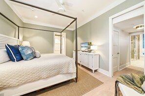 A four-poster bed with a white bedspread and blue pillows, a white dresser with a flat-screen TV, and a glass shower enclosure. - Oceanfront Jekyll Island • 3 King Bedrooms • Screened Porch • Walk to Dining (Jekyll Island)
