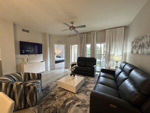 Ein modernes Wohnzimmer mit einem Flachbildfernseher, einem Deckenventilator, einem Sofa, einem Couchtisch und Blick auf ein Schlafzimmer durch eine geöffnete Tür.