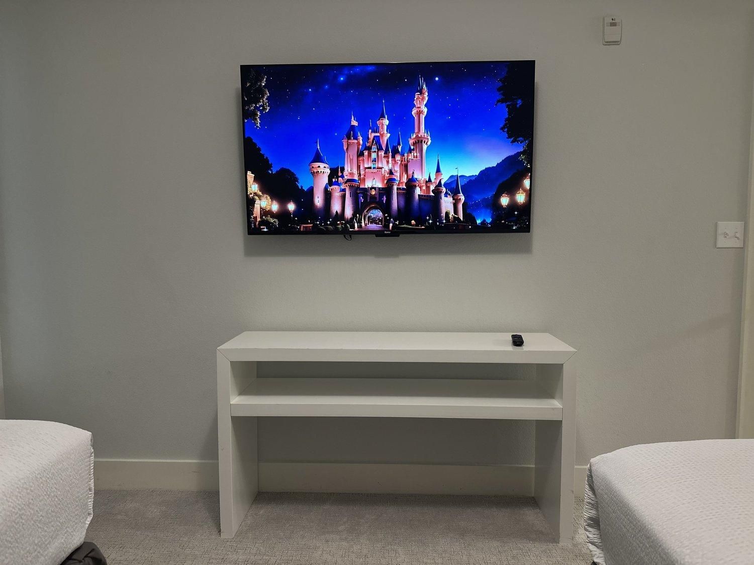 Une chambre à coucher avec un téléviseur fixé au mur, diffusant une scène de château, une console blanche et deux lits.