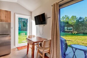A modern kitchen with a built-in refrigerator, a dining area with a wooden table and wicker chairs, a flat-screen TV mounted on the wall, and a view of a grassy outdoor area through a large window. - Glamping Pods at Tiffany's Estate | Hot Tub, Fire Pit, Pickleball, & Pond (Elk Park)