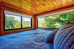A bedroom with a bed, two windows, and a view of trees. - Bear Pod at Tiffany's Estate | Hot Tub, Fire Pit, Pickleball, & Pond (Elk Park)