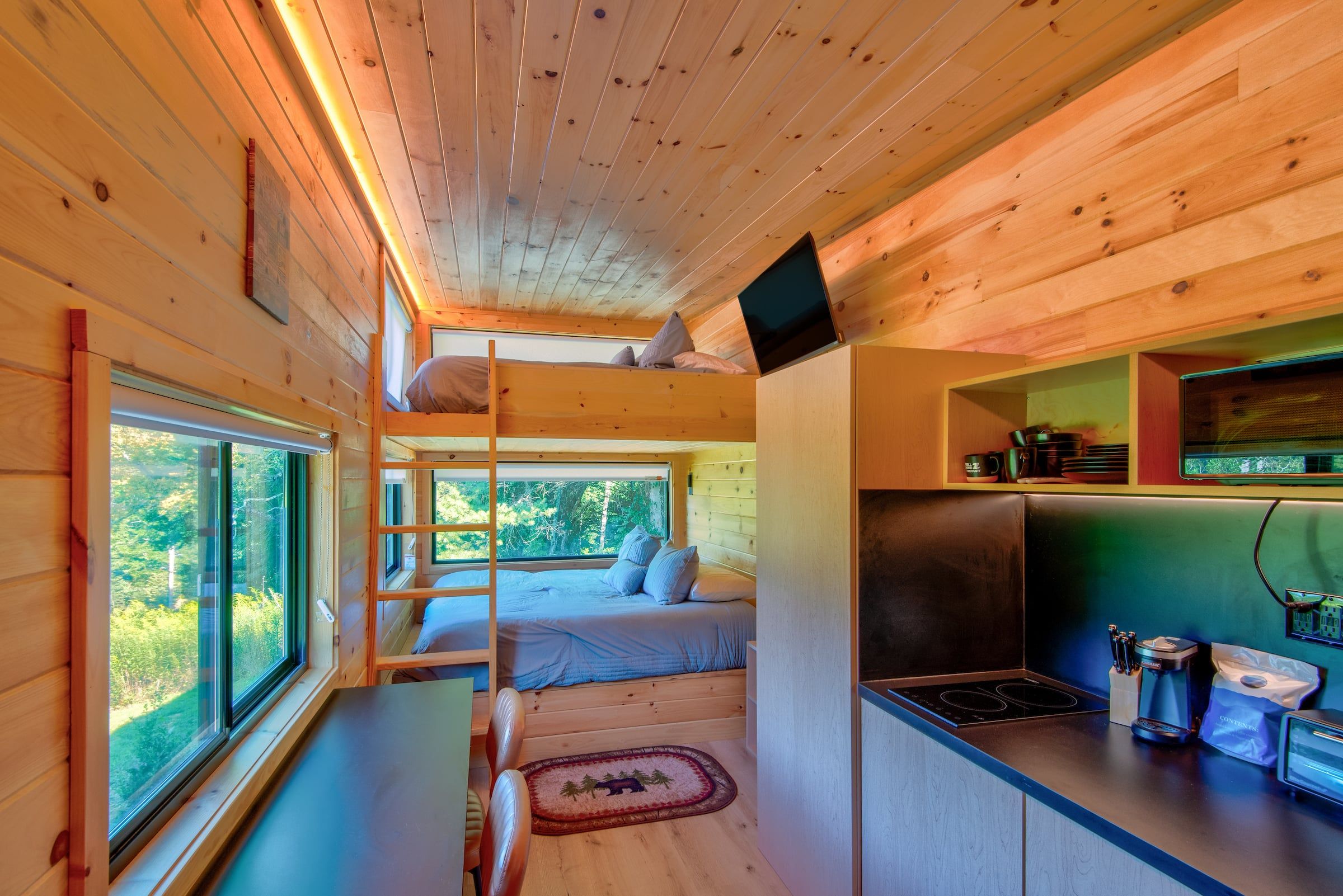 A compact cabin with a loft bed, a kitchenette, and a large window offering a view of the outdoors.