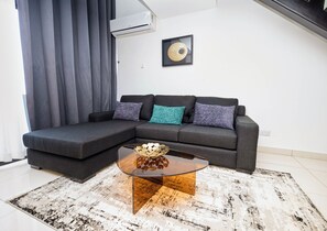 A modern living room with a sectional sofa, a glass coffee table, and a decorative wall piece. - Embassy Gardens Duplex by HDG (Accra)