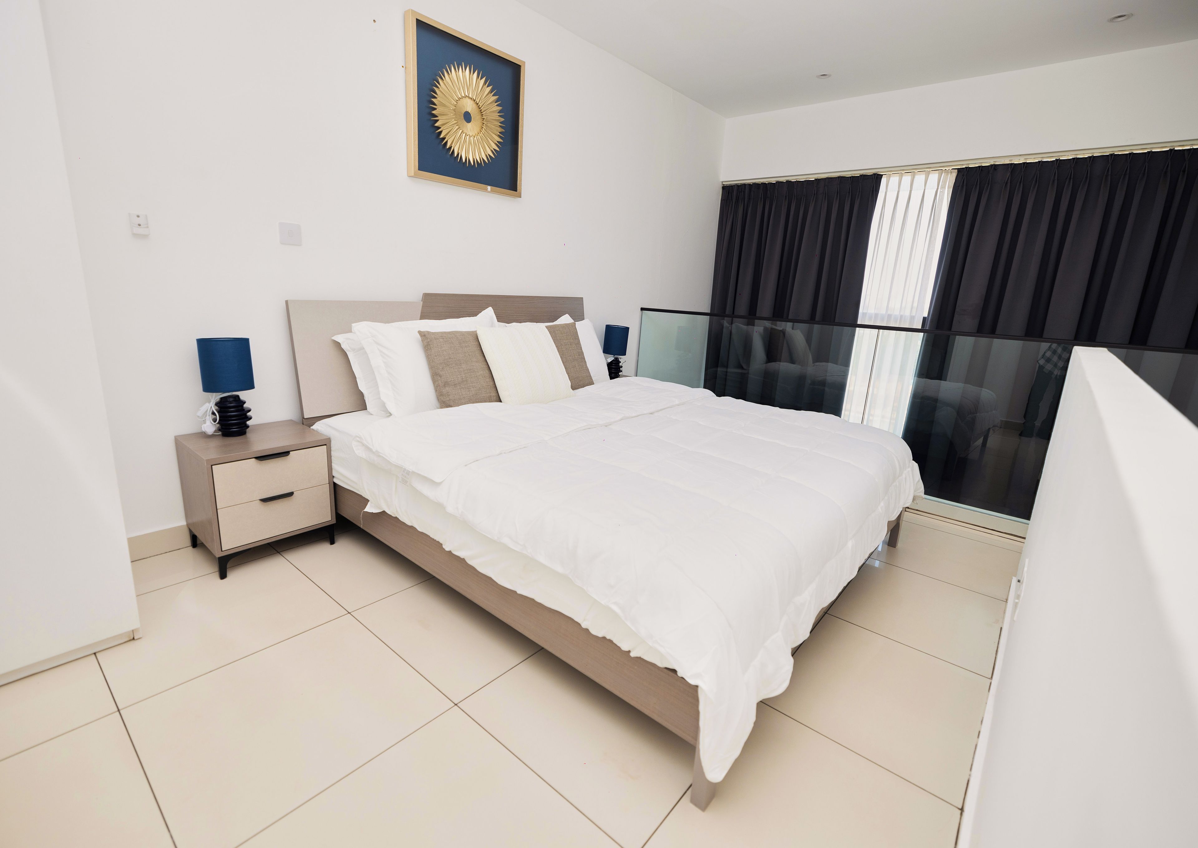 A modern bedroom with a bed, bedside tables, a nightstand, a wall-mounted lamp, and a large window with curtains.