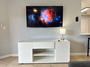 A flat-screen TV mounted on a wall above a white entertainment center with a remote control on it. - 1305 Stunning 3/3 Resort Condo Near Disney (Orlando)