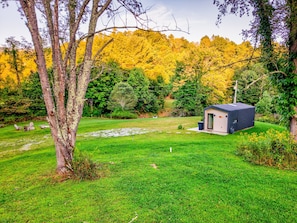 Eine kleine Holzhütte mit dunklem Dach und beigen Wänden, umgeben von einer grünen Wiese und Bäumen mit gelbem Herbstlaub.