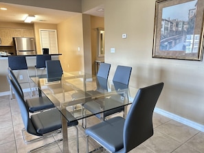 A modern kitchen with a stainless steel refrigerator, a dining area with a glass table and black chairs, and a framed cityscape painting on the wall. - 1405 Spacious 3/3 Resort Condo Near Disney (Orlando)