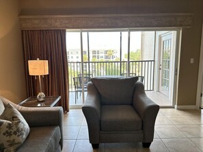 A hotel room with a sofa, a table with a lamp, a balcony with a view of buildings and greenery. - 1405 Spacious 3/3 Resort Condo Near Disney (Orlando)
