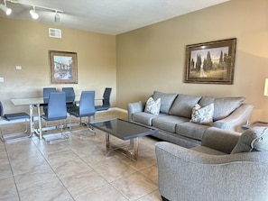 A living room with a grey sofa, a glass coffee table, and a dining area with chairs and a table. - 1405 Spacious 3/3 Resort Condo Near Disney (Orlando)