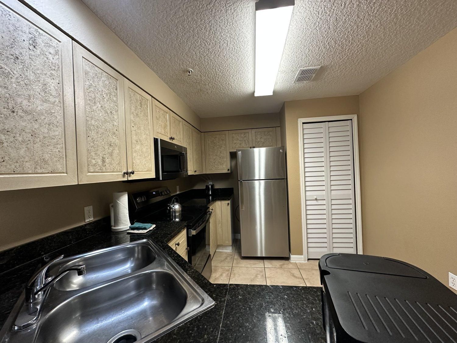 A kitchen with a stainless steel sink, a microwave, a refrigerator, and a black countertop.