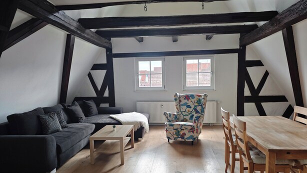 Quarto com sofá escuro, mesa de centro de madeira, poltrona com padrão floral e mesa de jantar com cadeiras.