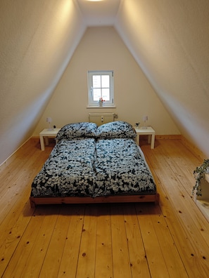 Un dormitorio con techo inclinado, una cama con ropa de cama floral, dos mesitas de noche, un ventanal con vistas y un radiador.