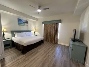 Ein Hotelzimmer mit einem Bett, zwei Nachttischen mit Lampen, einem Deckenventilator, einem Fernseher auf einem Schrank und einem Fenster mit Vorhängen.