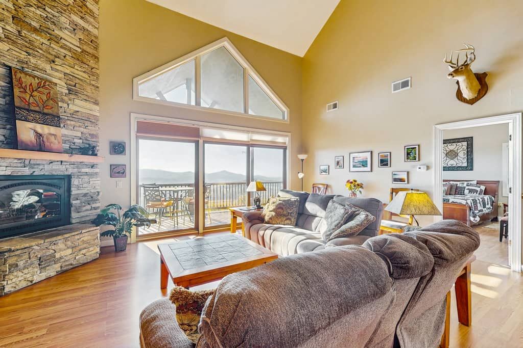 A spacious living room with a stone fireplace, large windows, and a high ceiling.