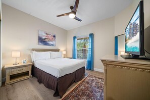 Habitación de hotel con una cama, un escritorio, un televisor, un ventilador de techo, una ventana con cortinas y un cuadro en la pared.