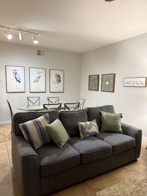 A living room with a gray sofa, a glass dining table, and wall art including bird paintings. - 2305 Spacious 3/3 Resort Condo Near Disney (Orlando)