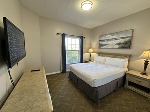 Quarto de hotel com cama, escrivaninha, televisão, janela com cortinas, um quadro na parede e abajur.