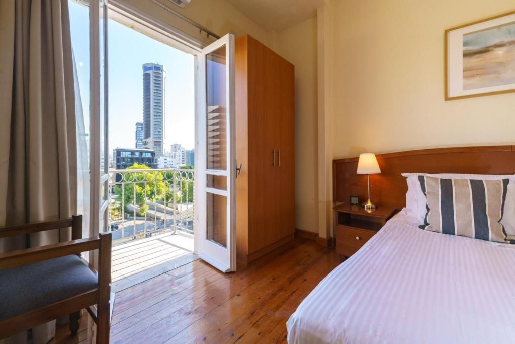 Habitación de hotel con cama, mesita de noche, armario de madera, balcón y vistas a edificios, además de una silla.