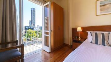 A hotel room with a bed, a nightstand, a wooden wardrobe, a balcony with a view of buildings, and a chair.