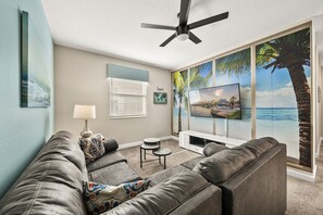 A living room with a grey sofa, a small table, a TV, and a large window with a view of palm trees and a beach. - 2779 Oxymoron Way (Kissimmee)