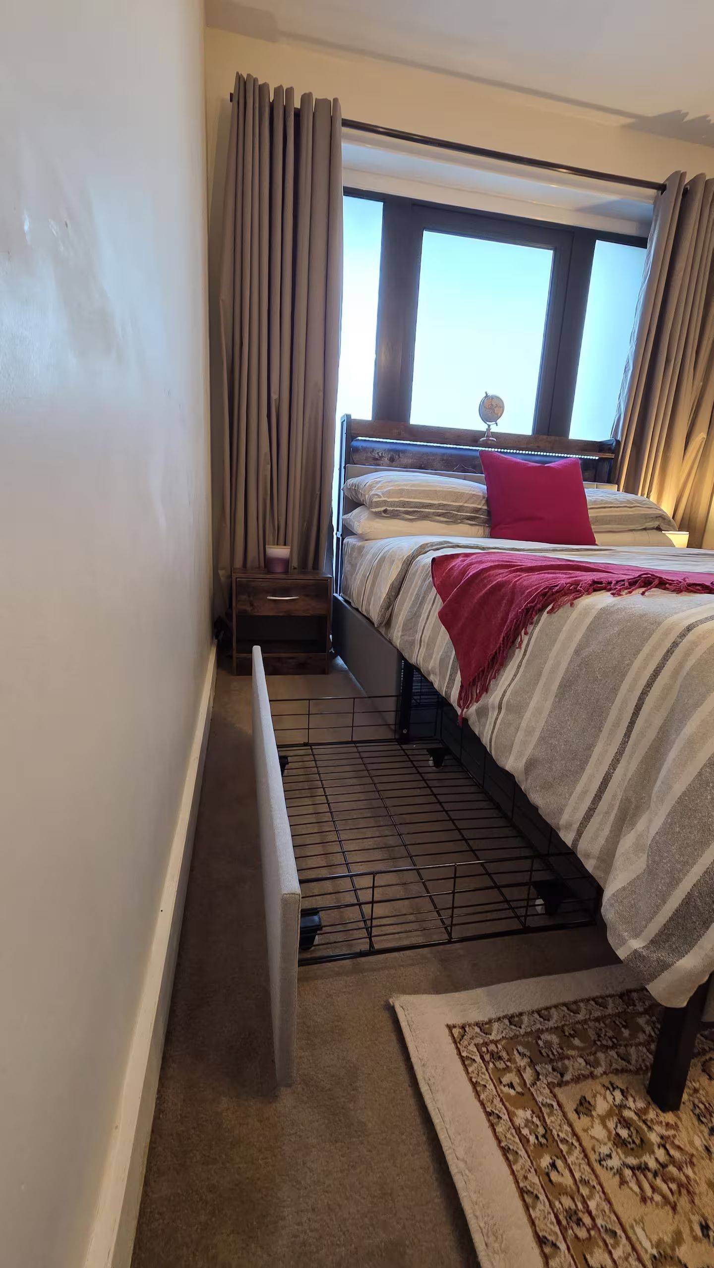 Un dormitorio con cama, radiador, ventana con cortinas y una alfombra estampada.