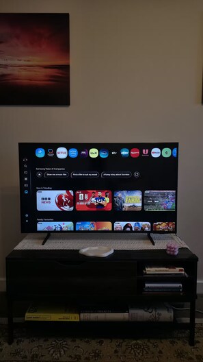 Ein Fernseher auf einem Standfuß mit Auswahl an Streaming-Diensten und einem darunterliegenden Bücherregal.