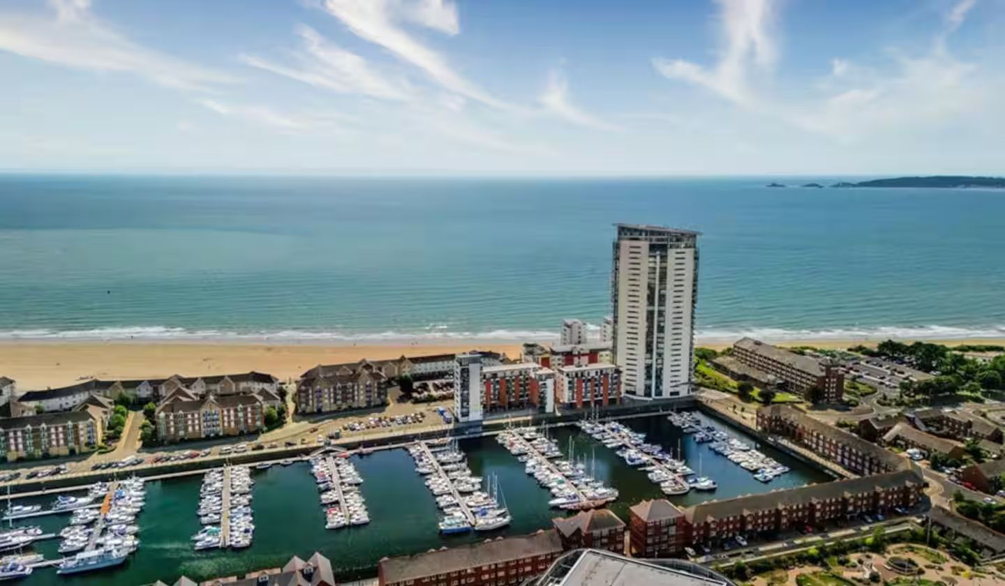 Ein Küstenhafen mit mehreren angedockten Booten, einem hohen Hotelgebäude und einem Strand im Hintergrund.