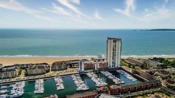Una marina costiera con diverse imbarcazioni ormeggiate, un alto edificio di un hotel e una spiaggia sullo sfondo.