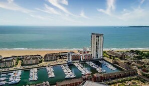 Un puerto deportivo costero con varias embarcaciones amarradas, un alto edificio de hotel y una playa al fondo.