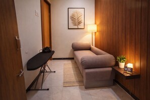 A compact room with a sofa, a small table with a lamp, a potted plant, and a clock displaying 12:25. - Hotel Shree Kripa (Ujjain)
