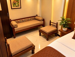 A hotel room with a bed, a sofa, a chair, and a nightstand with a plant. - Hotel Shree Kripa (Ujjain)