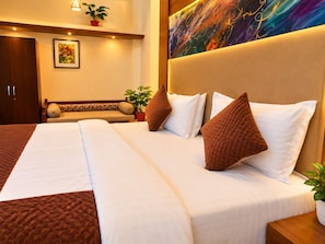 Habitación de hotel con una cama grande, cabecera con un cuadro, una banca con cojines y una planta en maceta.