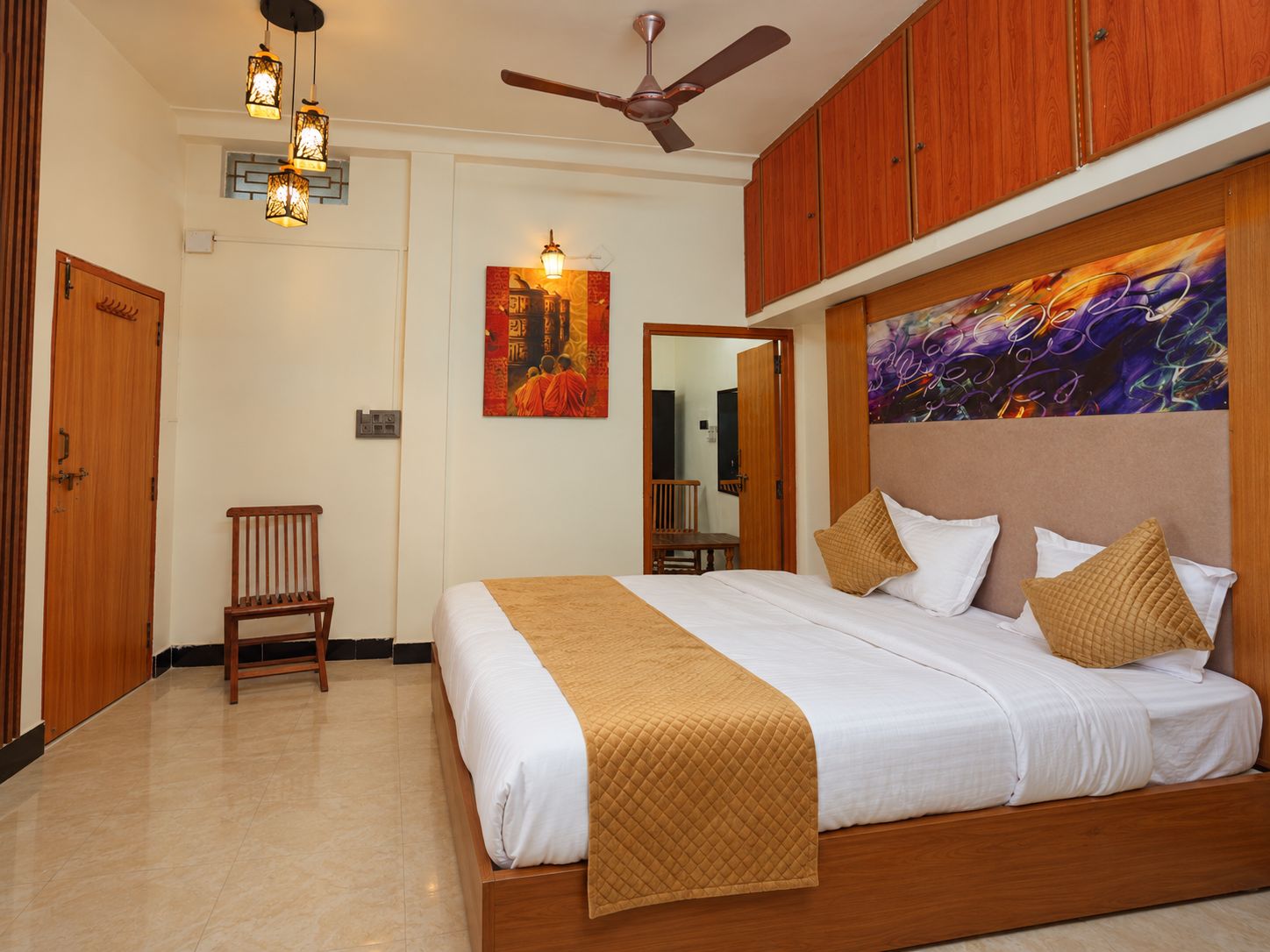A hotel room with a bed, a ceiling fan, wooden cabinets, and artwork on the walls.