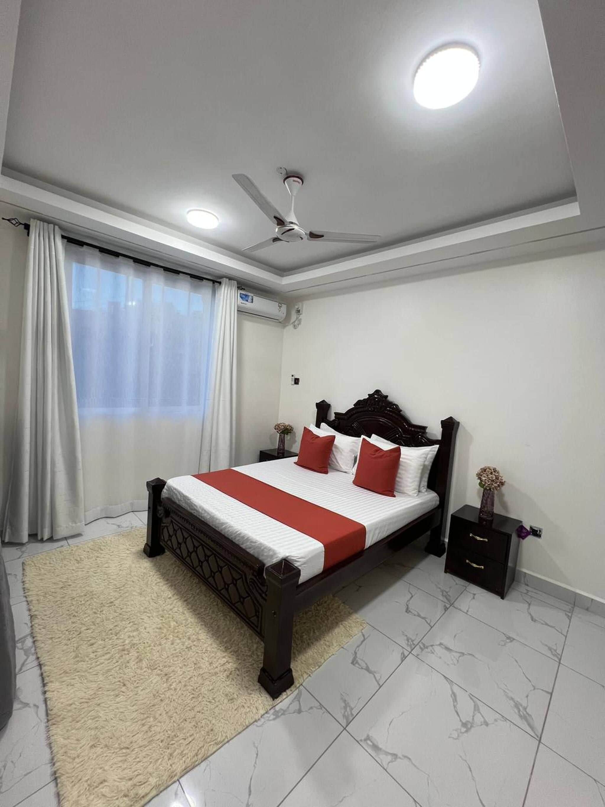 Une chambre à coucher avec un lit, une table de chevet, un ventilateur de plafond et une fenêtre avec des rideaux.