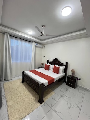 Un dormitorio con cama, mesita de noche, ventilador de techo y ventana con cortinas.