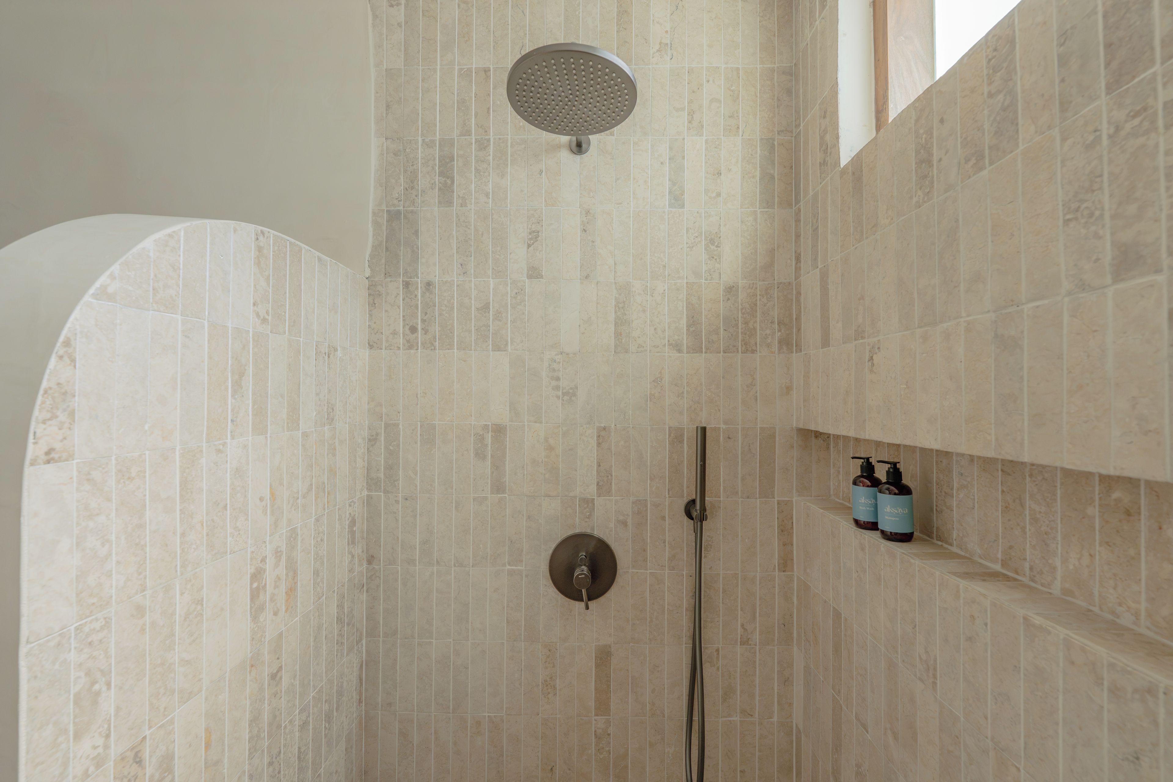 A shower area with a rain showerhead, a handheld showerhead, and a shelf with two bottles of liquid.