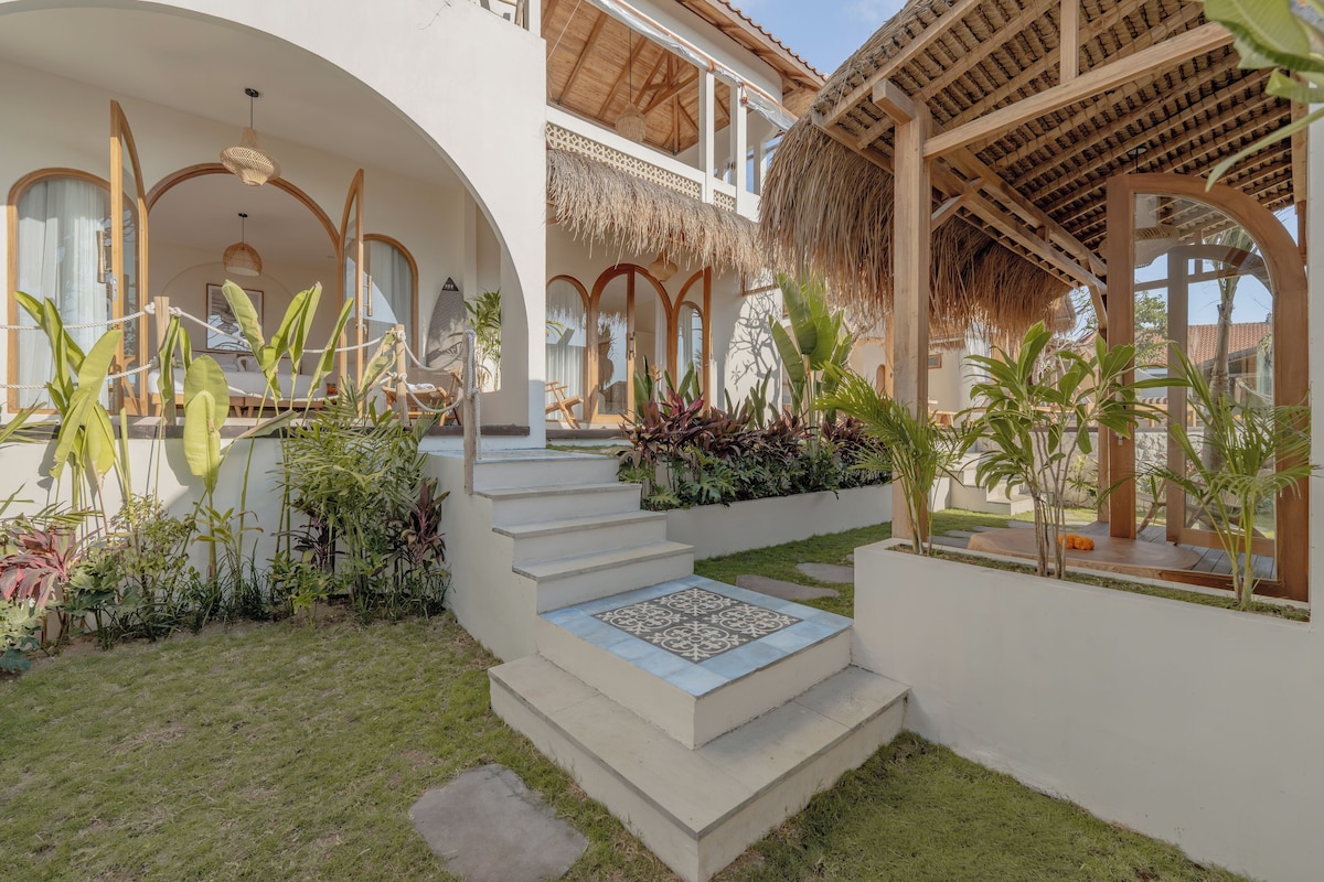 Un espace extérieur moderne doté d’un escalier menant à un coin salon abrité, entouré d’une végétation luxuriante et surmonté d’une structure à toit de chaume.