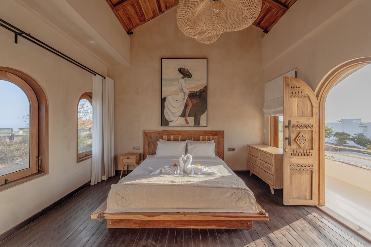 Une chambre à coucher avec un grand lit, une tête de lit en bois, un tableau, une armoire en bois et une vue sur l’extérieur par une porte ouverte.