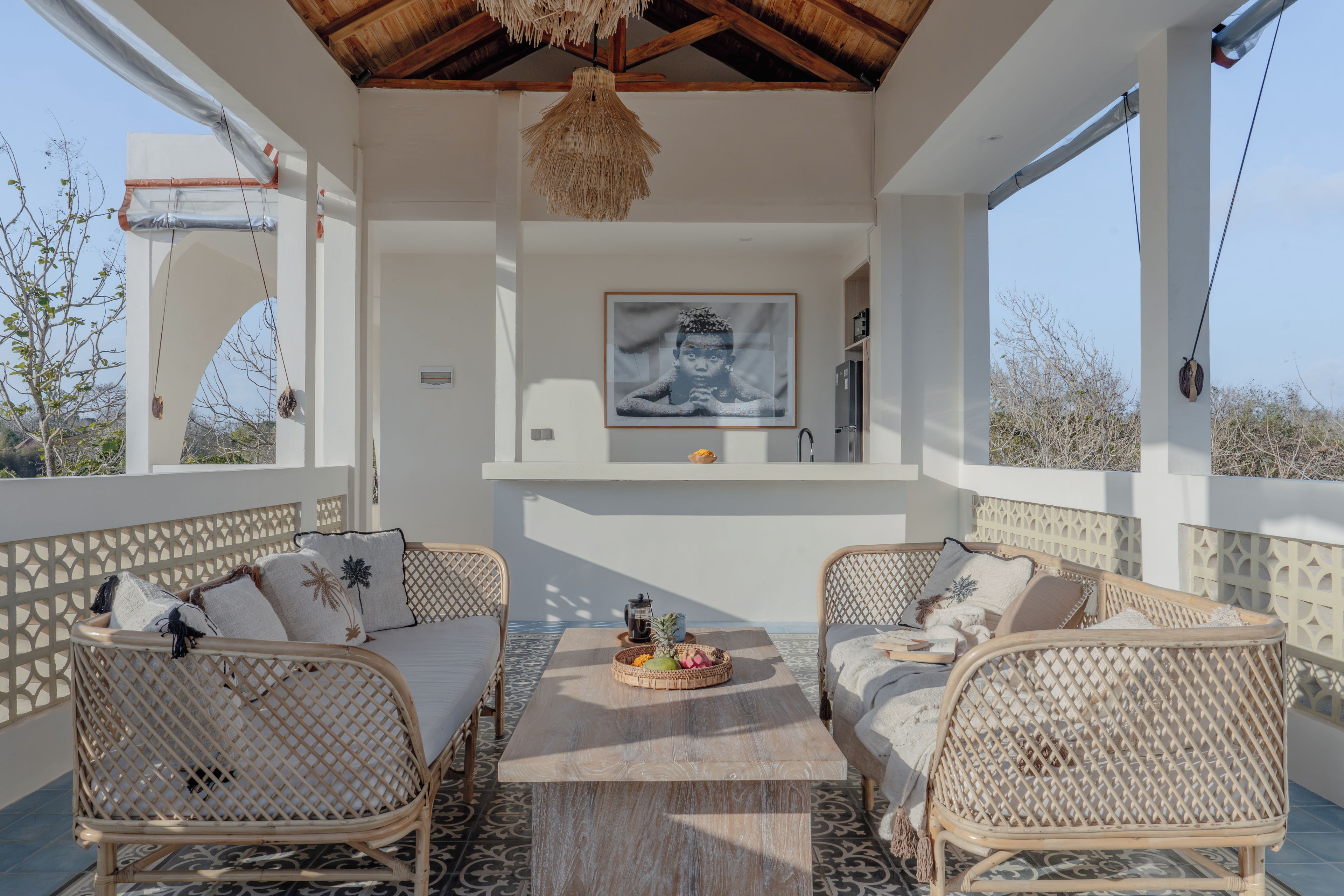 A sunlit outdoor seating area with wicker furniture, a central table, and a framed artwork on the wall.