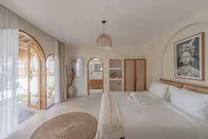 Une chambre avec un grand lit, une armoire en bois, une salle de bain avec un miroir et une vue sur l’extérieur par une porte ouverte.