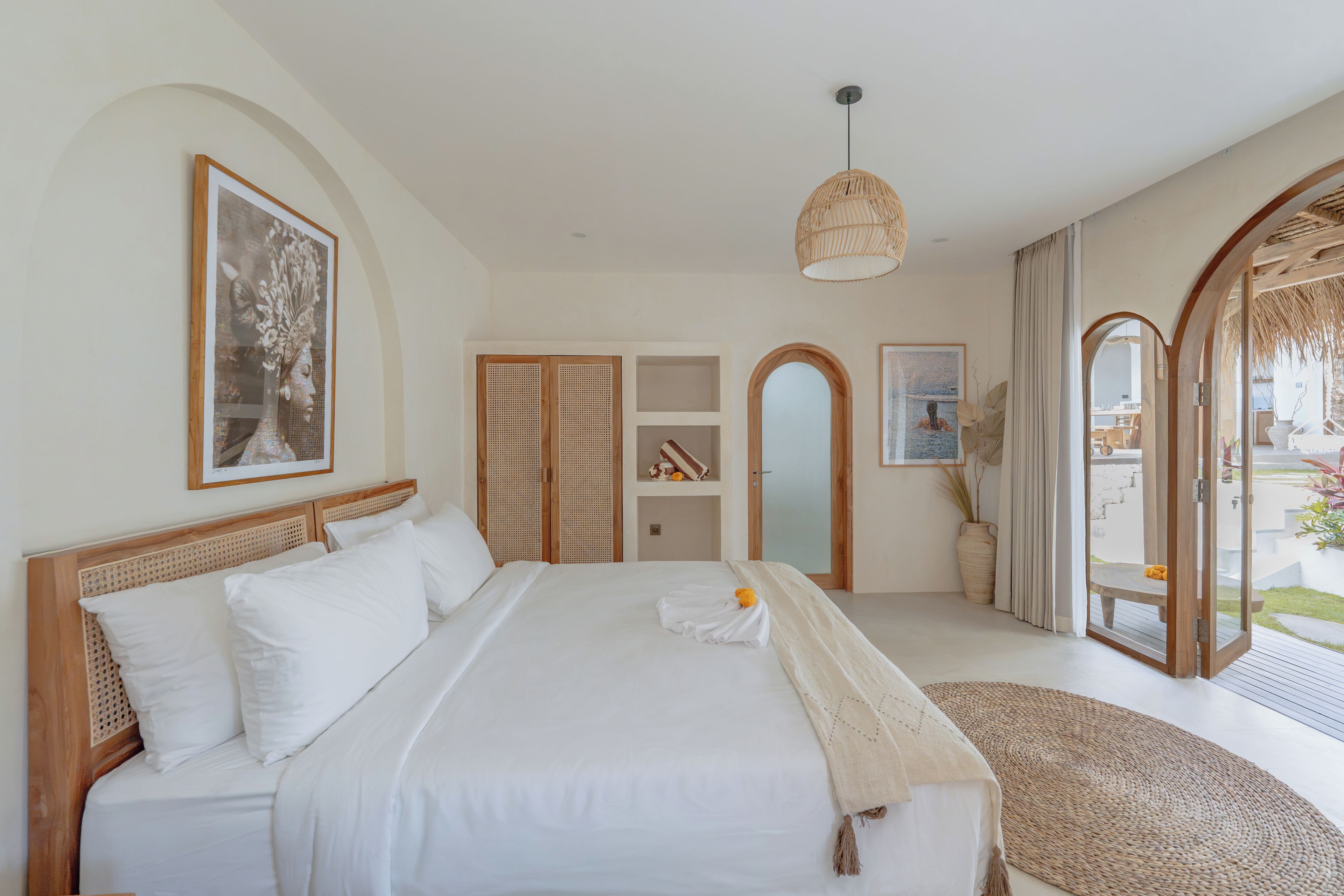 A bedroom with a large bed, a wooden headboard, a woven floor mat, and a view of the outside.