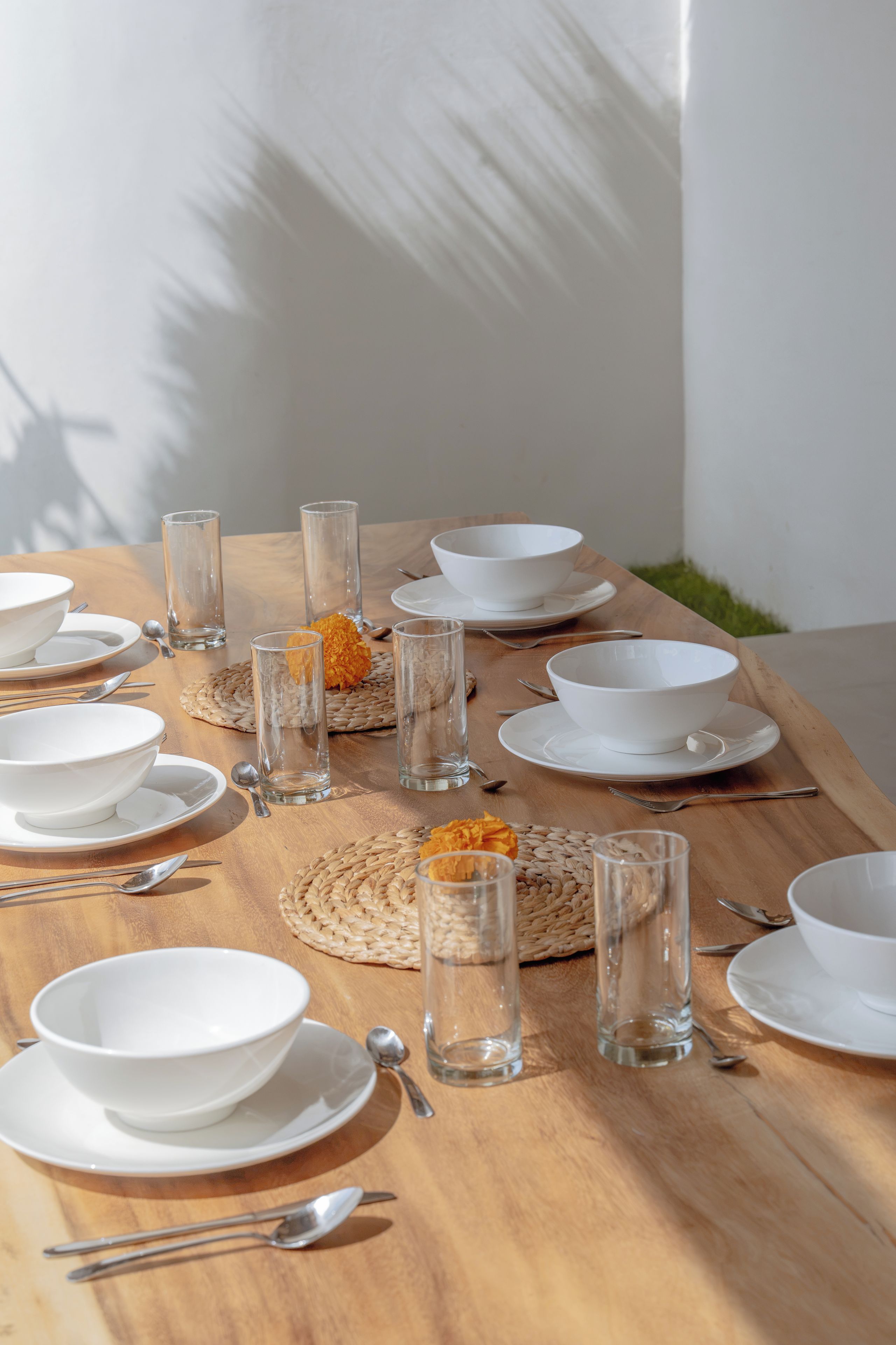 Una mesa de comedor dispuesta con cuencos blancos, vasos y cubiertos, con un protector de mesa tejido y un florero con flores.